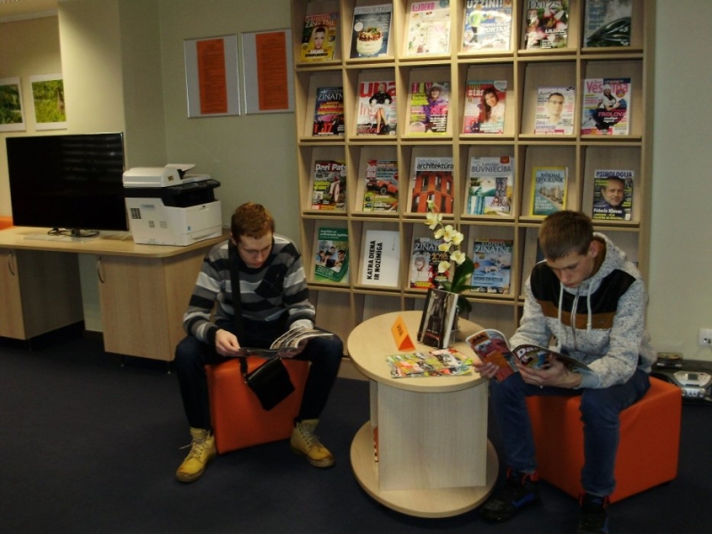 Skolas audzēkņi lasa bibliotēkā pie žurnālu stenda, sēž uz pufiem
